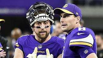 Two Vikings players in purple jerseys talk on sideline during game.