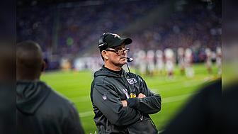 Coach stands on Vikings sideline during NFL game, arms crossed.