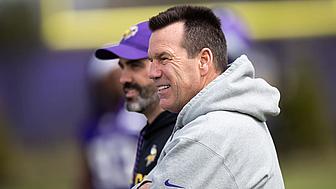 Man wearing purple Vikings cap and gray hoodie smiles during game.