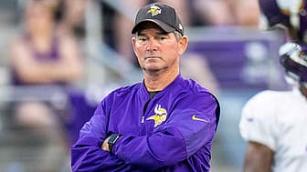 Coach in purple Vikings jacket with arms crossed on sideline.
