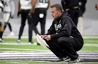 Coach crouches on sideline holding clipboard during NFL game.