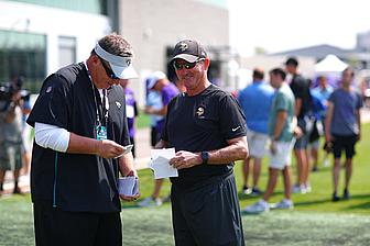 Two coaches review clipboard during outdoor Vikings training camp practice session.
