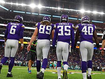 Four Vikings players walking on field wearing purple jerseys and white pants.