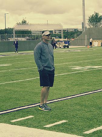 Coach standing on football field during practice with players in background.