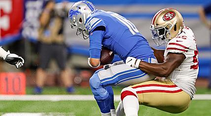 Lions player in blue tackled by 49ers defender in red and gold uniform.