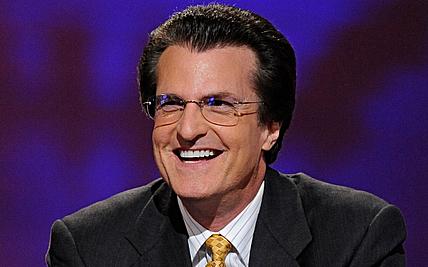Man wearing glasses and dark suit smiling against purple background.