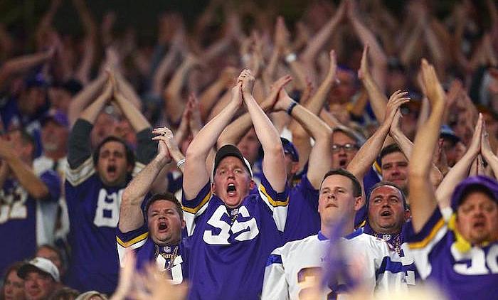 Fans in purple Vikings jerseys cheering with arms raised at stadium.
