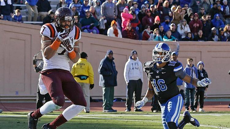 Football player in maroon jersey catches ball while defender in blue pursues.