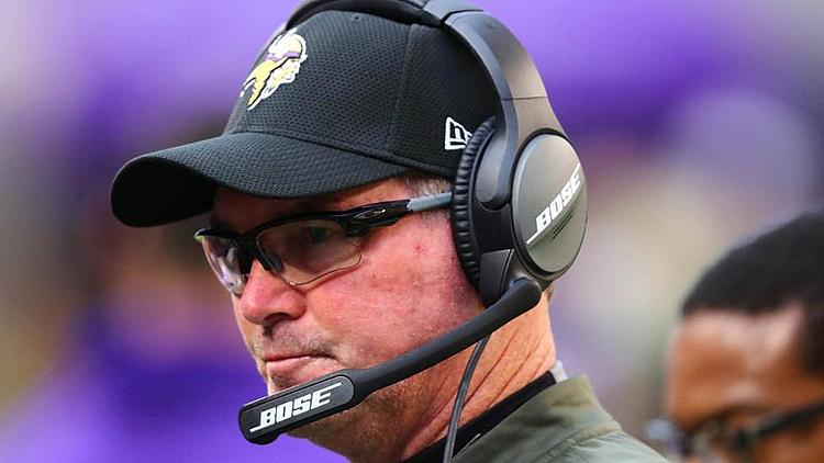 Coach wearing Vikings cap and headset during game on sideline.