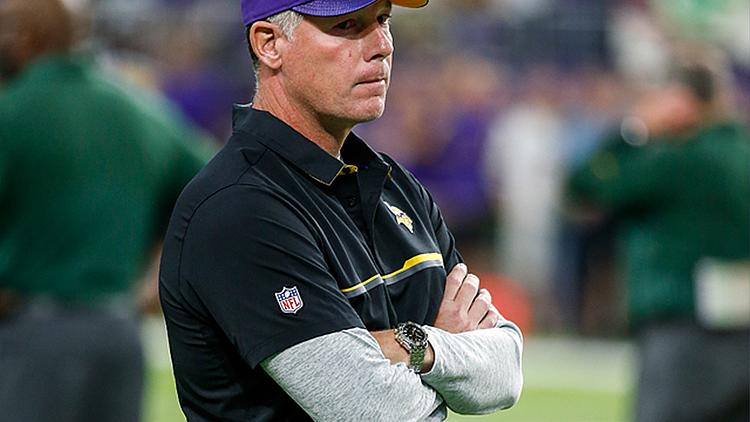 Coach wearing purple Vikings cap observes game from sideline.