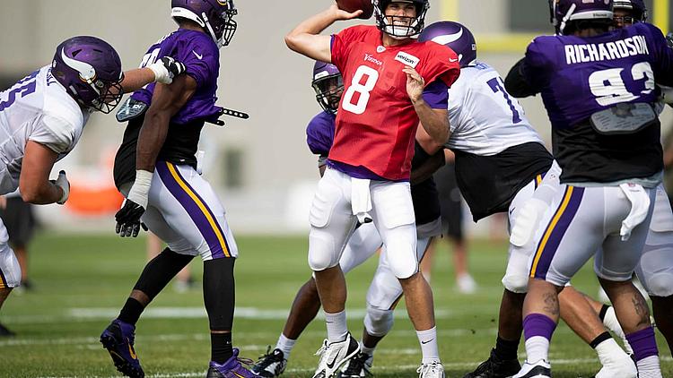 Vikings quarterback in red jersey during practice drill with purple jersey defenders.