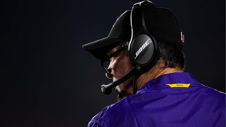 Coach wearing purple Vikings jacket and headset on sideline.
