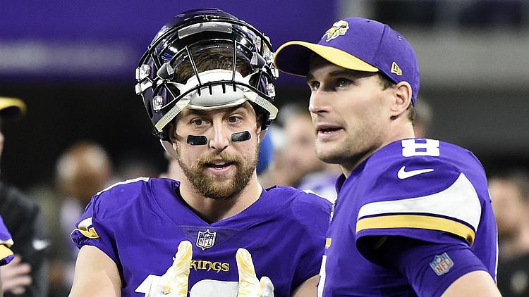 Two Vikings players in purple jerseys talk on sideline during game.