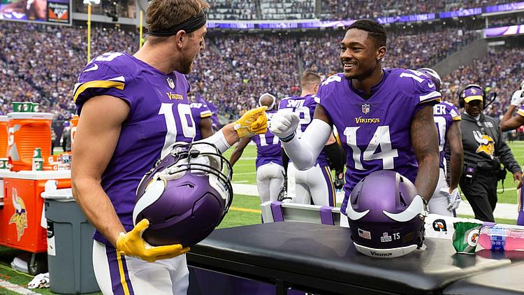Two Vikings players in purple jerseys celebrate on sideline during game.