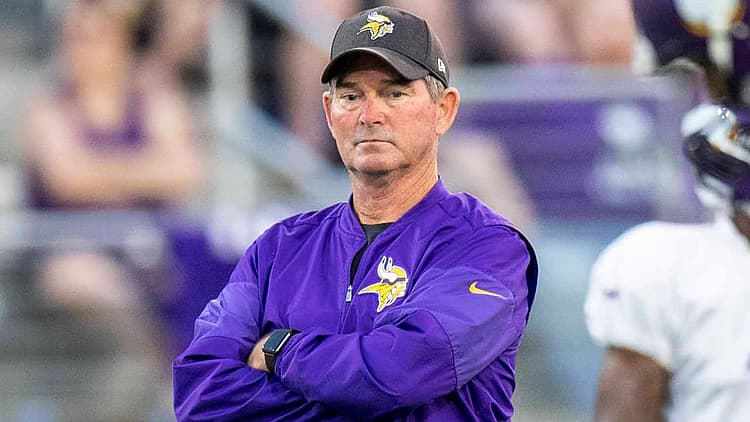 Coach in purple Vikings jacket with arms crossed on sideline.