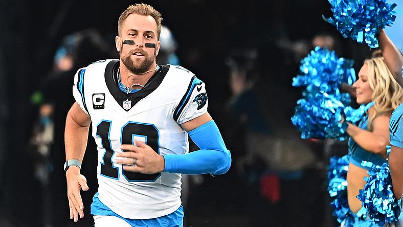 Football player in white and blue jersey running onto field with cheerleaders.