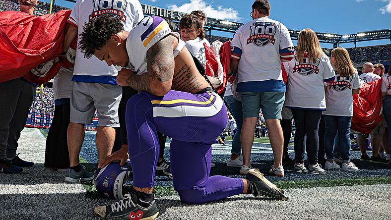 Player in purple Vikings uniform kneels on field during stadium event with fans.